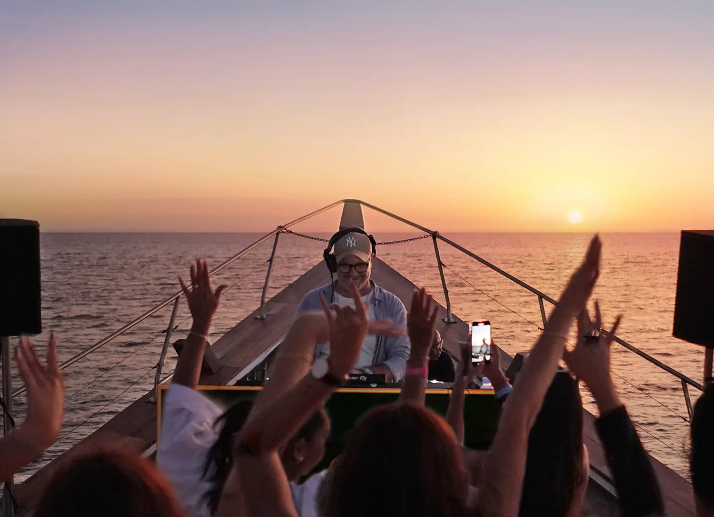 DJ in headphones performing at the bow as guests dance with raised hands at sunset