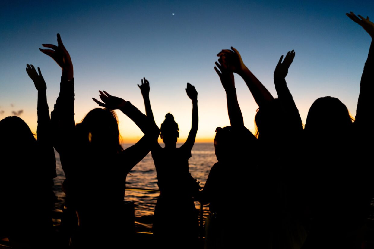 Guests dancing with raised arms silhouetted against an ocean sunset