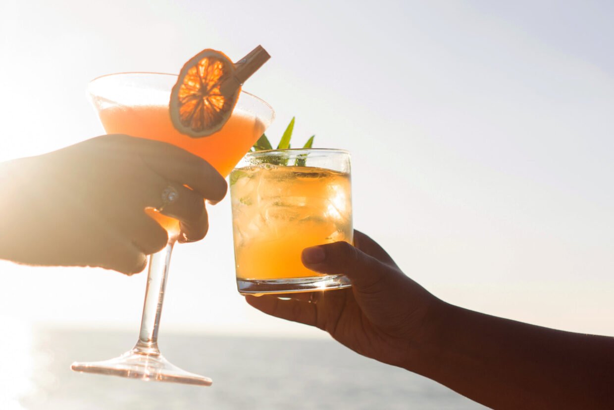 Two cocktails clinked in a toast against a bright sunset sea