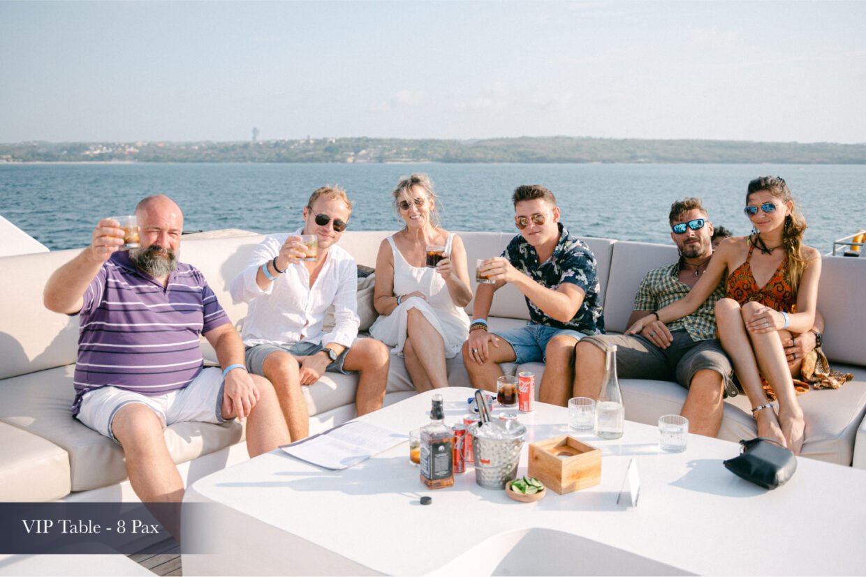 Six guests raising drinks at a VIP table with the Nusa Dua coastline behind