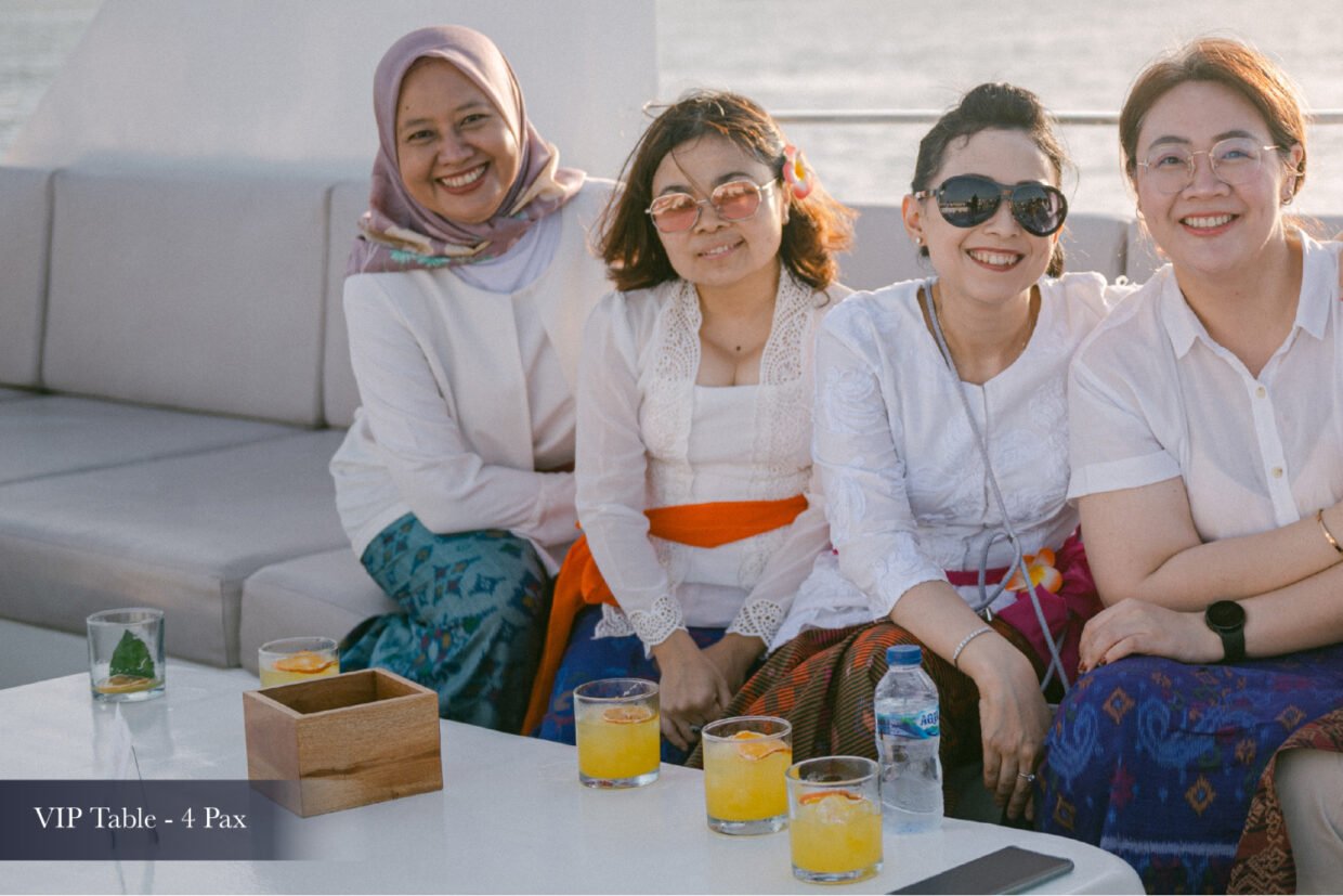 Four women in white smiling at a VIP table with cocktails at sunset