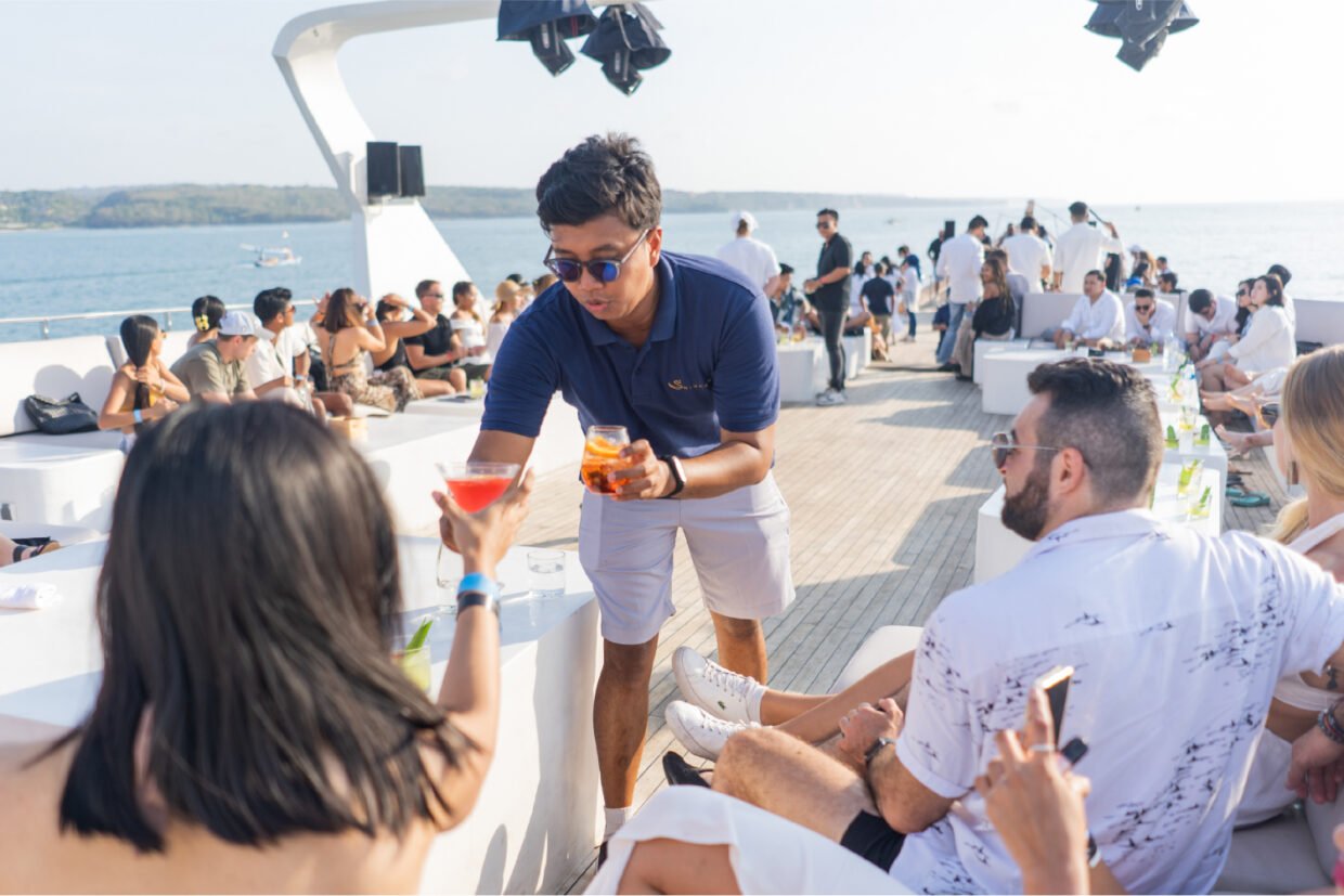 Host serving cocktails to seated guests on deck with Nusa Dua behind