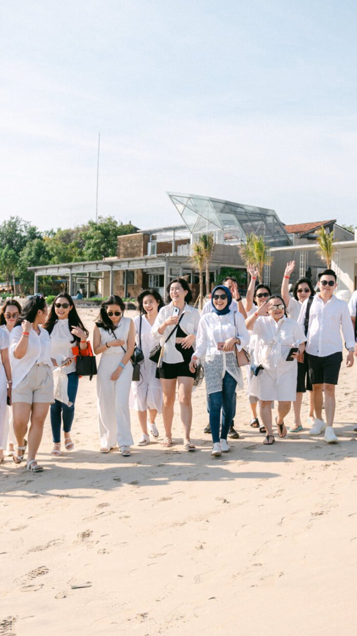Group of guests in white walking the beach past Benoa waterfront buildings