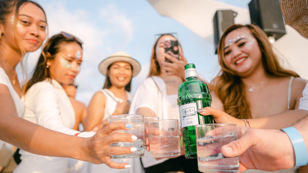 Women with festival face glitter toasting drinks around a gin bottle on deck