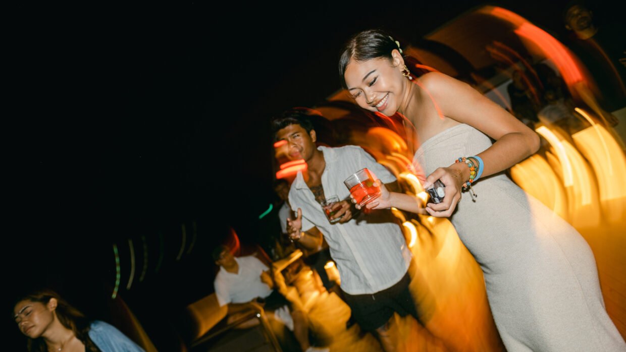 Smiling woman in a strapless dress holding a drink amid party lights on deck