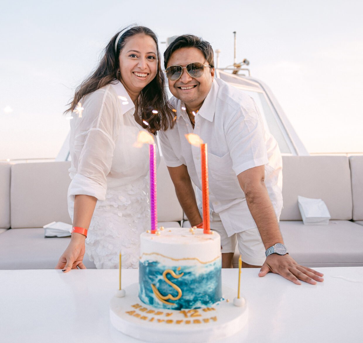 Couple in white posing behind a Shivanna anniversary cake on the sunlit deck