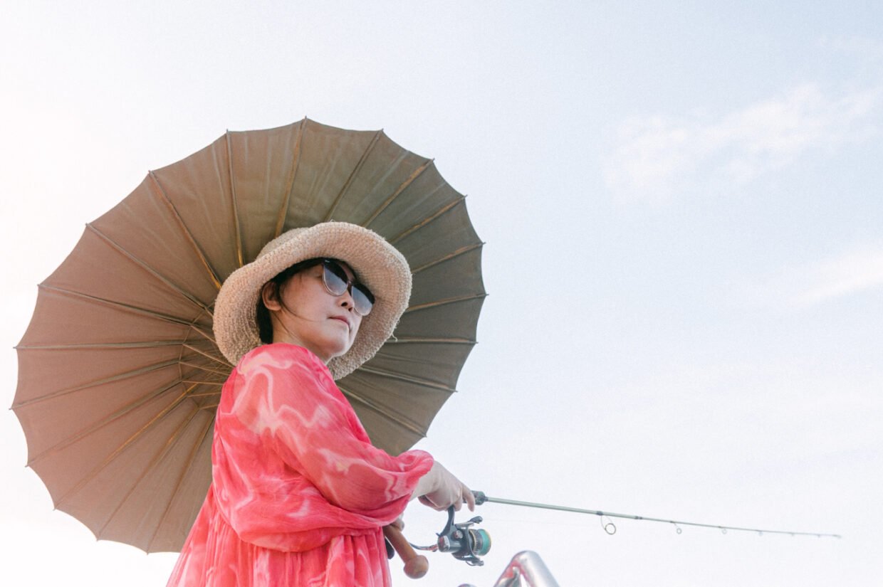 Woman in a straw hat and pink dress fishing under a parasol against a bright sky