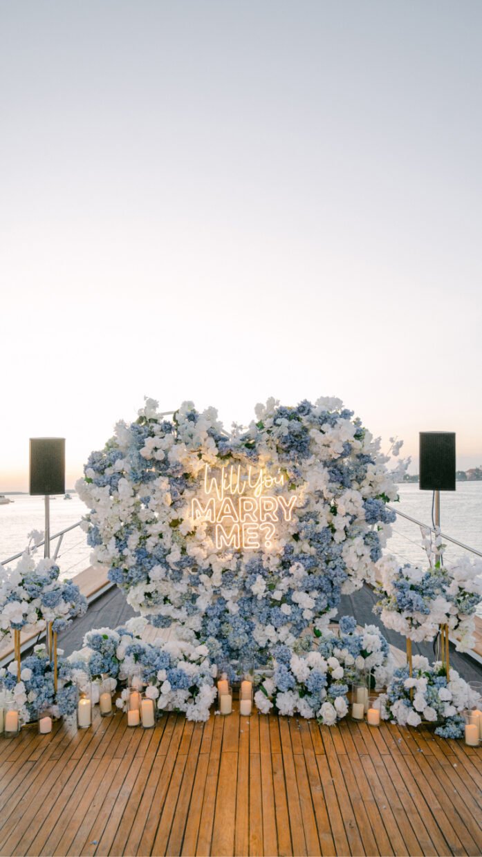 Heart-shaped floral display with a "Will You Marry Me?" neon sign on deck at dusk