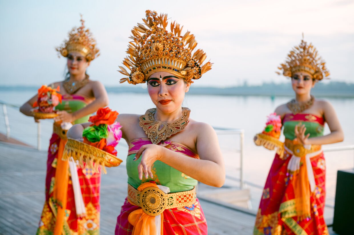 Three Balinese dancers in gilded headdresses and bright costumes perform on deck at dusk