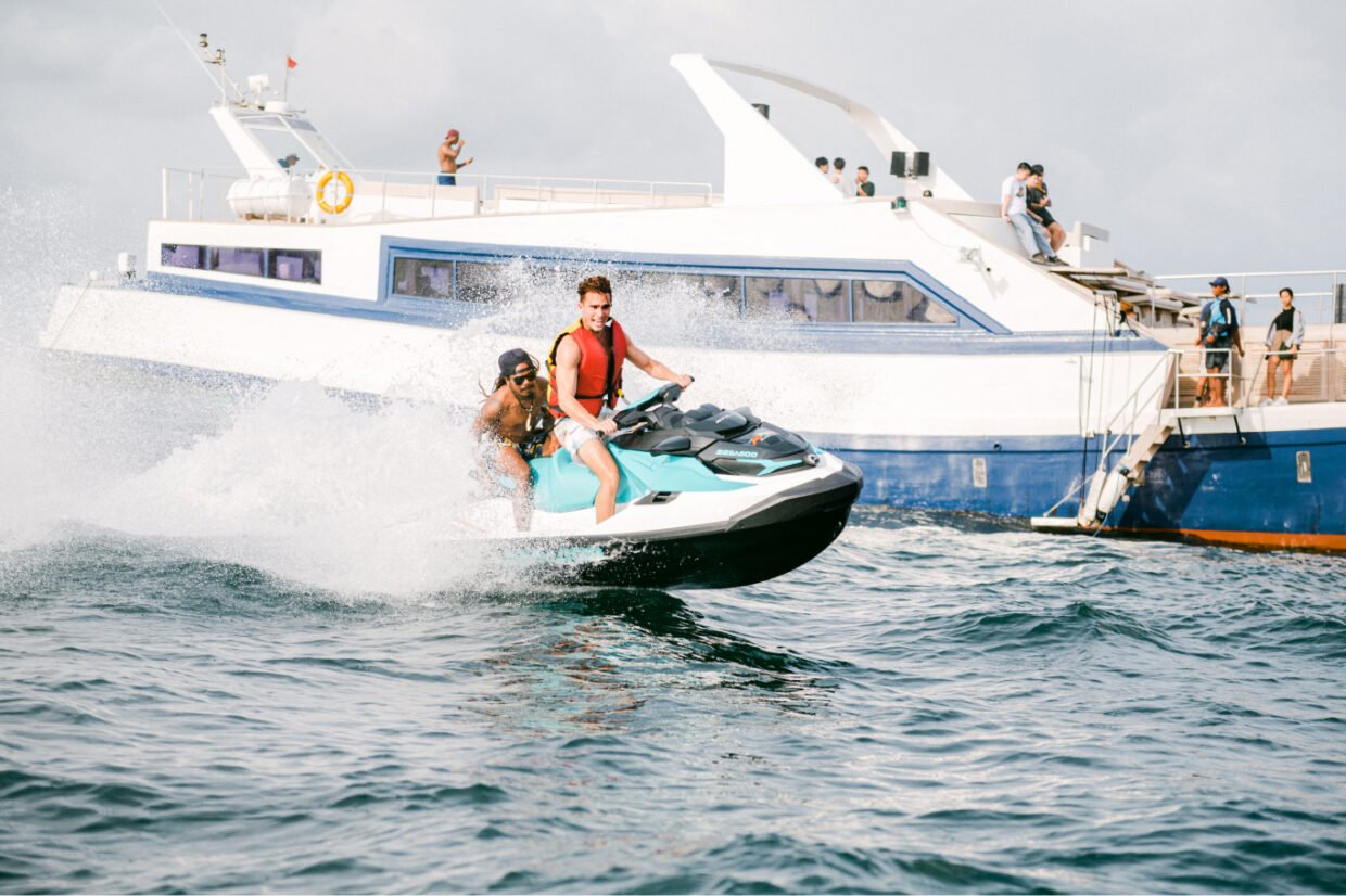 Two men ride a jet ski past a moored cruise boat with onlookers