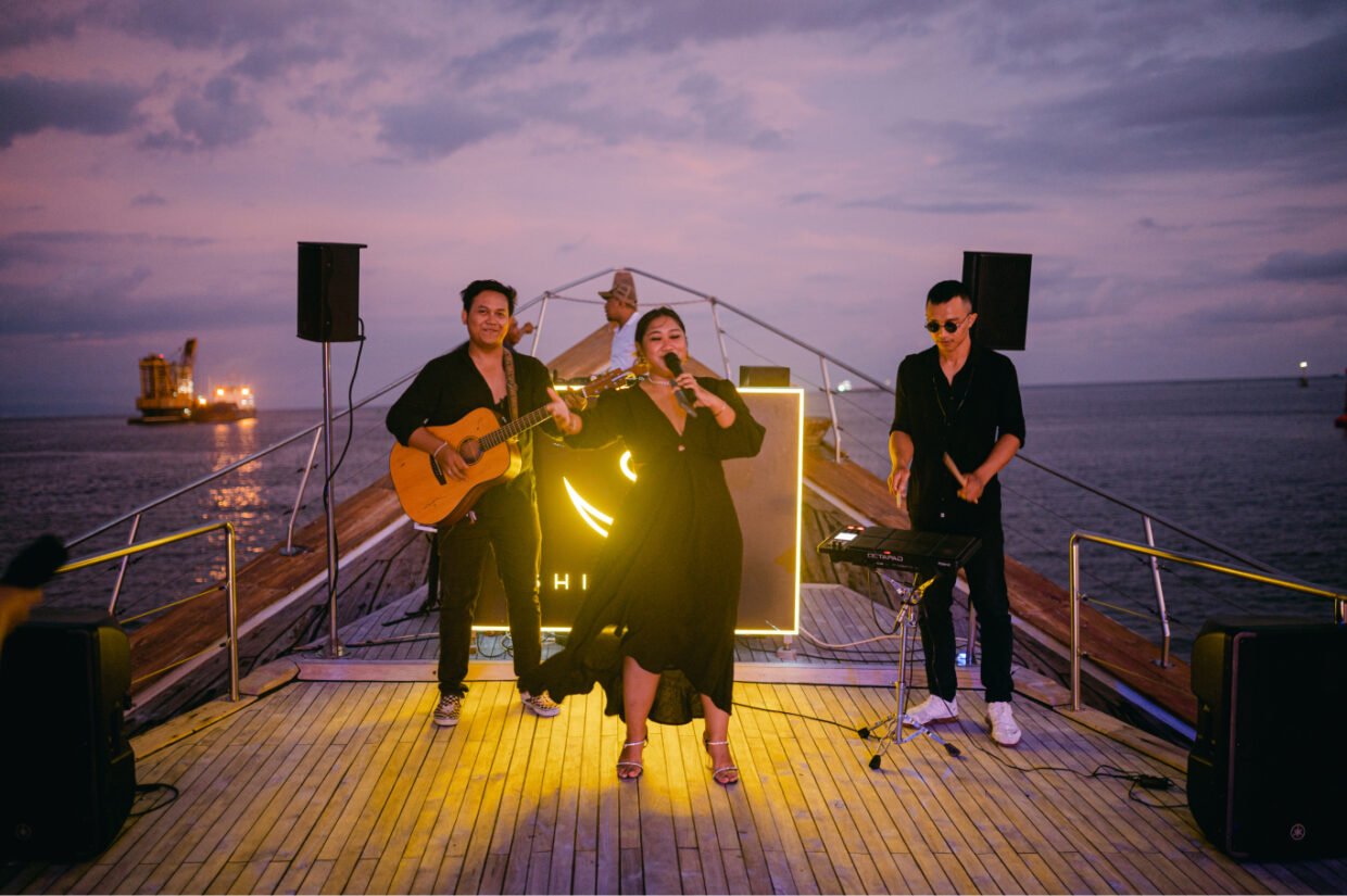 Three-piece band with a singer in black performing on the deck bow at sunset