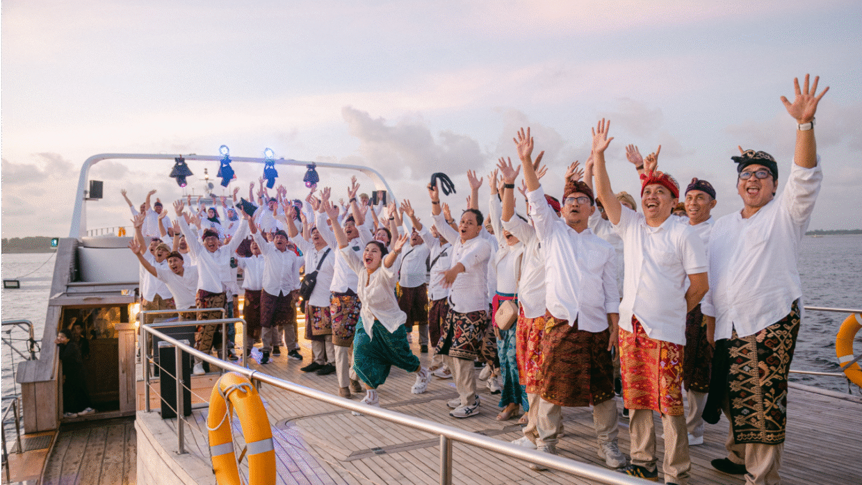 Large group in white shirts and Balinese sarongs cheering with raised arms on deck at dusk