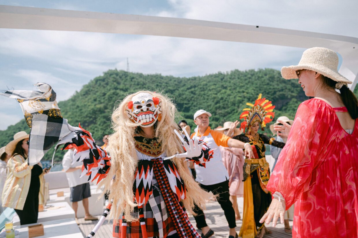 Costumed Balinese performer with guests dancing on deck against green Nusa Dua hills
