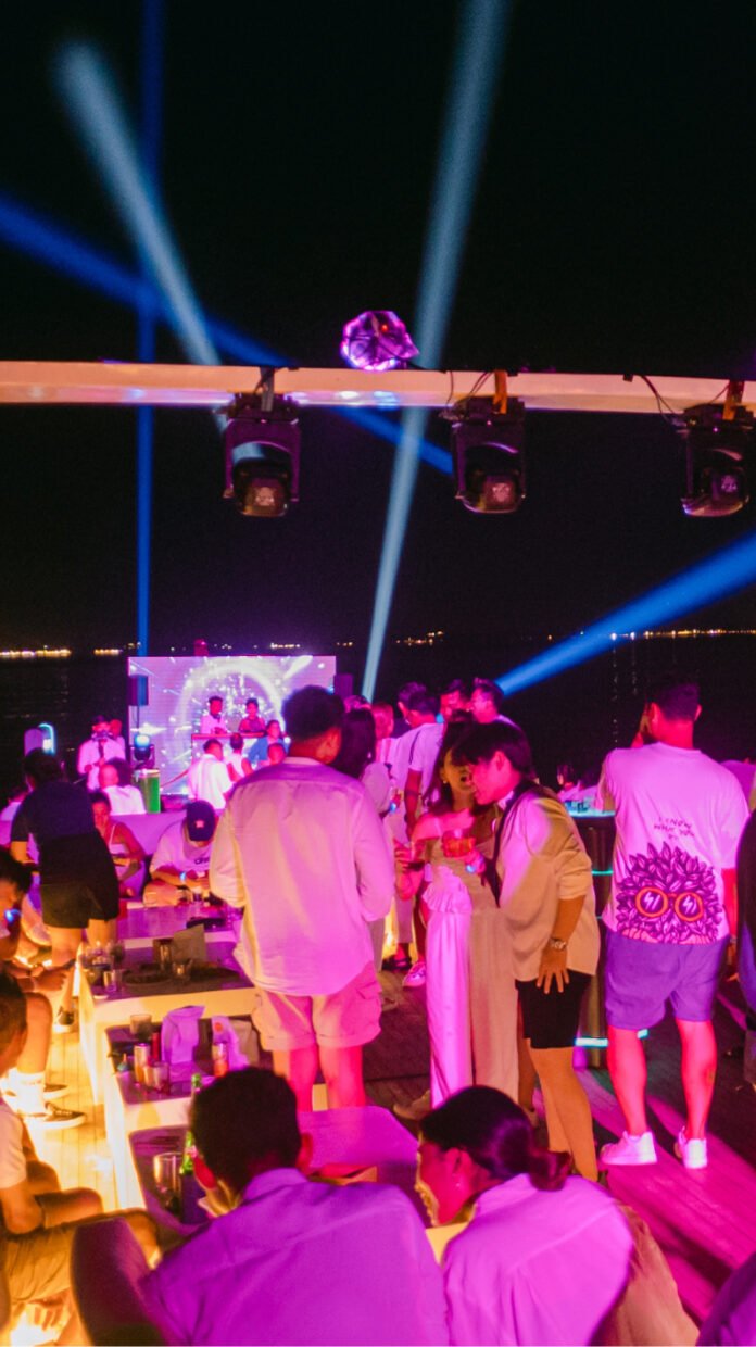 Crowded night deck party under blue stage lights with DJ and screen