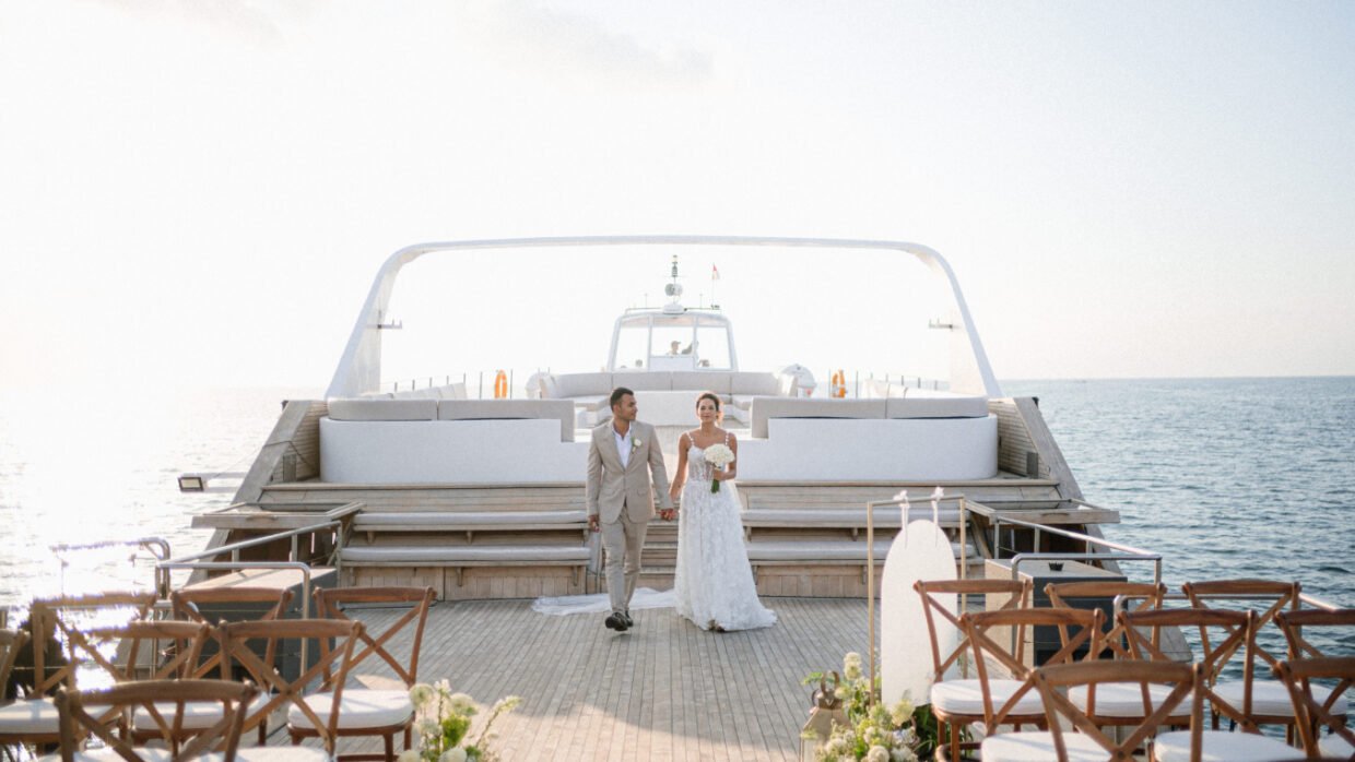 Friends in white celebrating with arms raised behind a cocktail-laden table on deck
