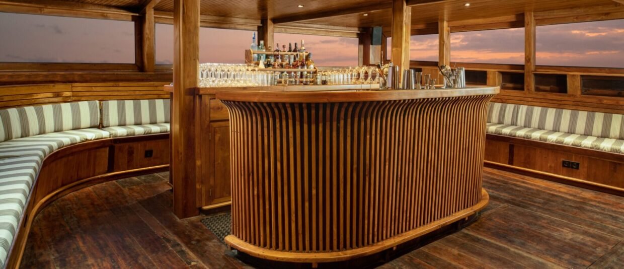 Curved timber-slatted bar stocked with spirits and glassware on deck at dusk