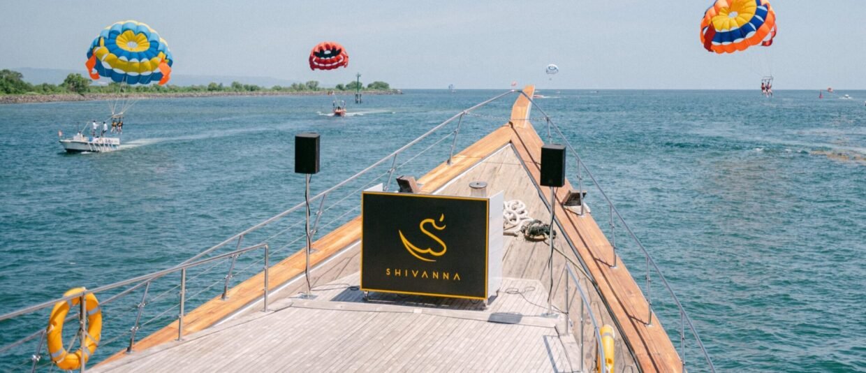 The Shivanna yacht bow with gold swan sign and teak deck, parasailers over the Nusa Dua water