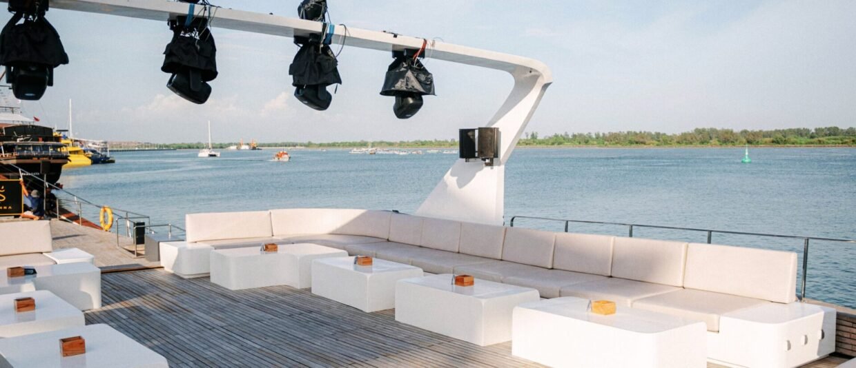 White-cushioned upper deck lounge under a row of stage lights, harbour and moored boats behind