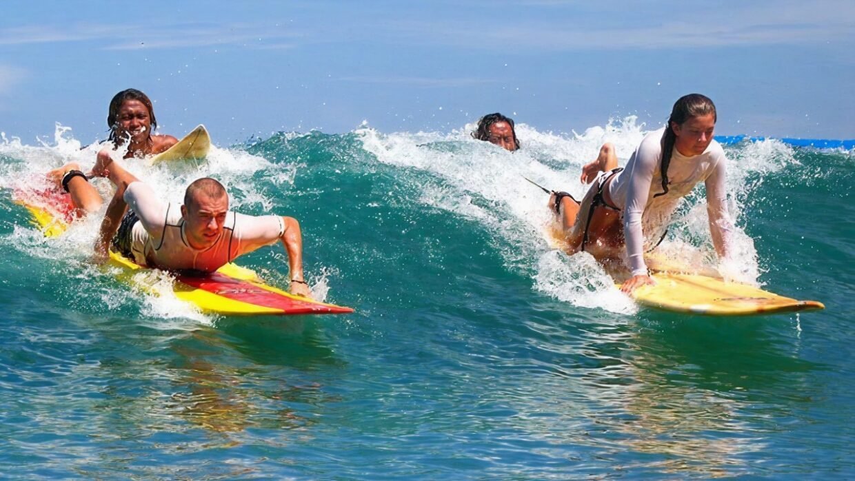Four surfers riding waves in clear blue ocean, two in the front and two behind, enjoying dynamic surfing on brightly colored boards under a sunny sky.