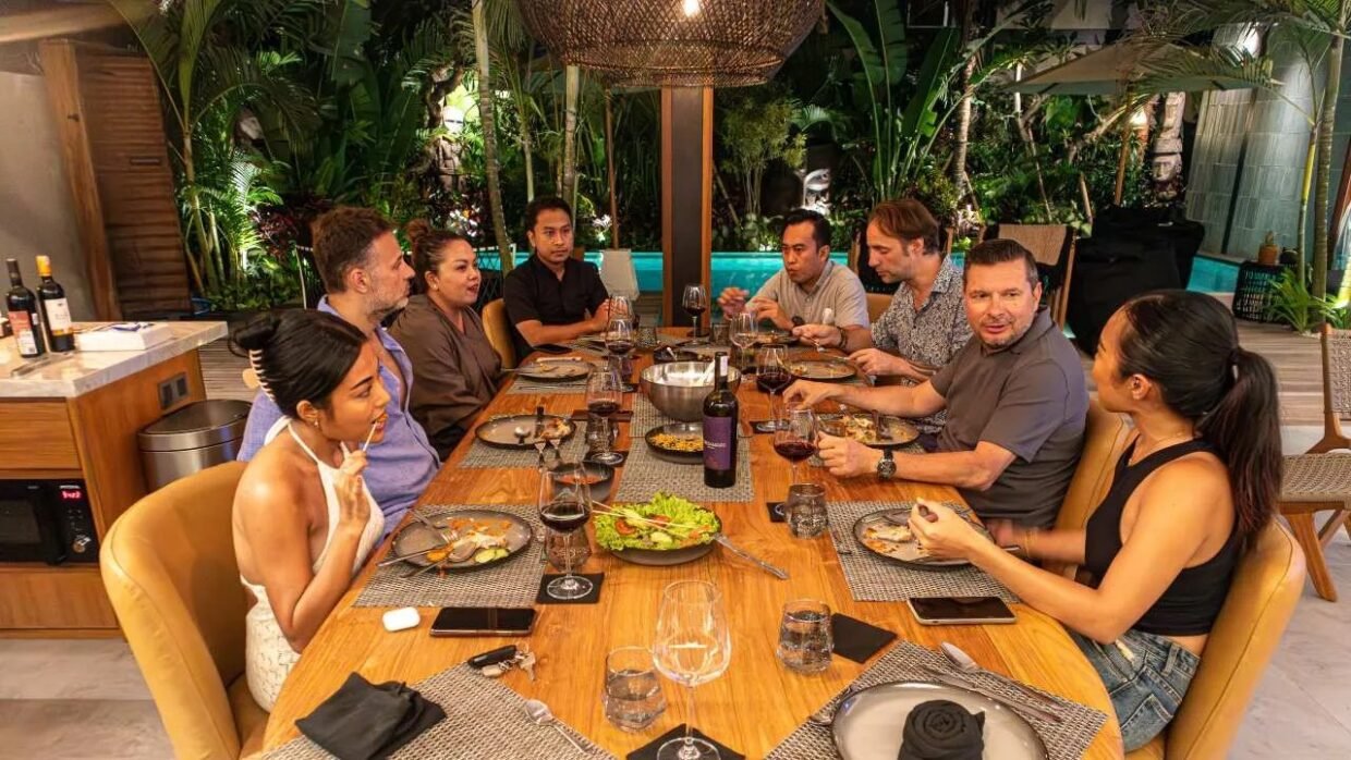 People enjoying a dinner gathering around a wooden table in a tropical outdoor setting