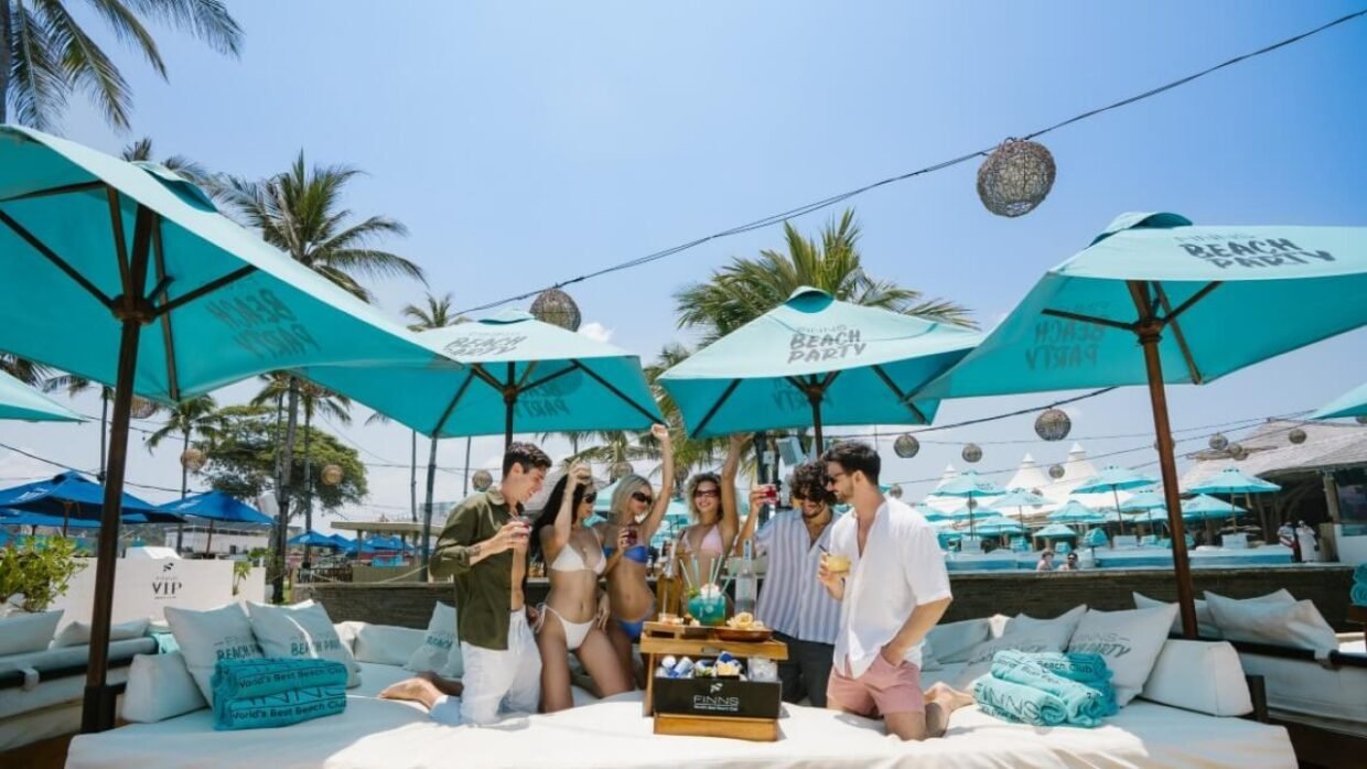 Friends enjoying drinks on a lounge at a Bali beach club under turquoise umbrellas.